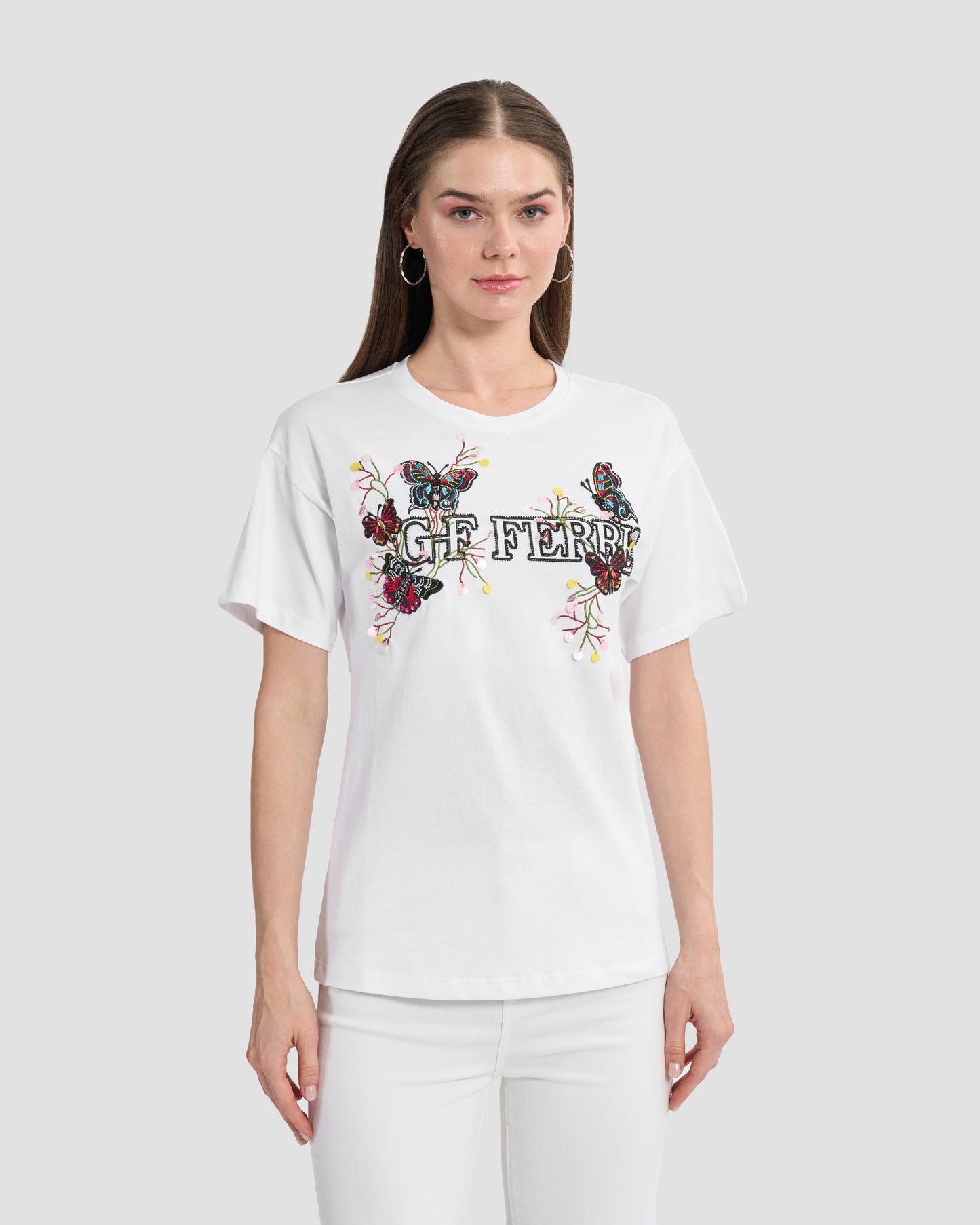 A woman wearing a white t-shirt with butterfly patches to the front, beaded Ferré branding, and multi-toned sequins paired with a white jeans.