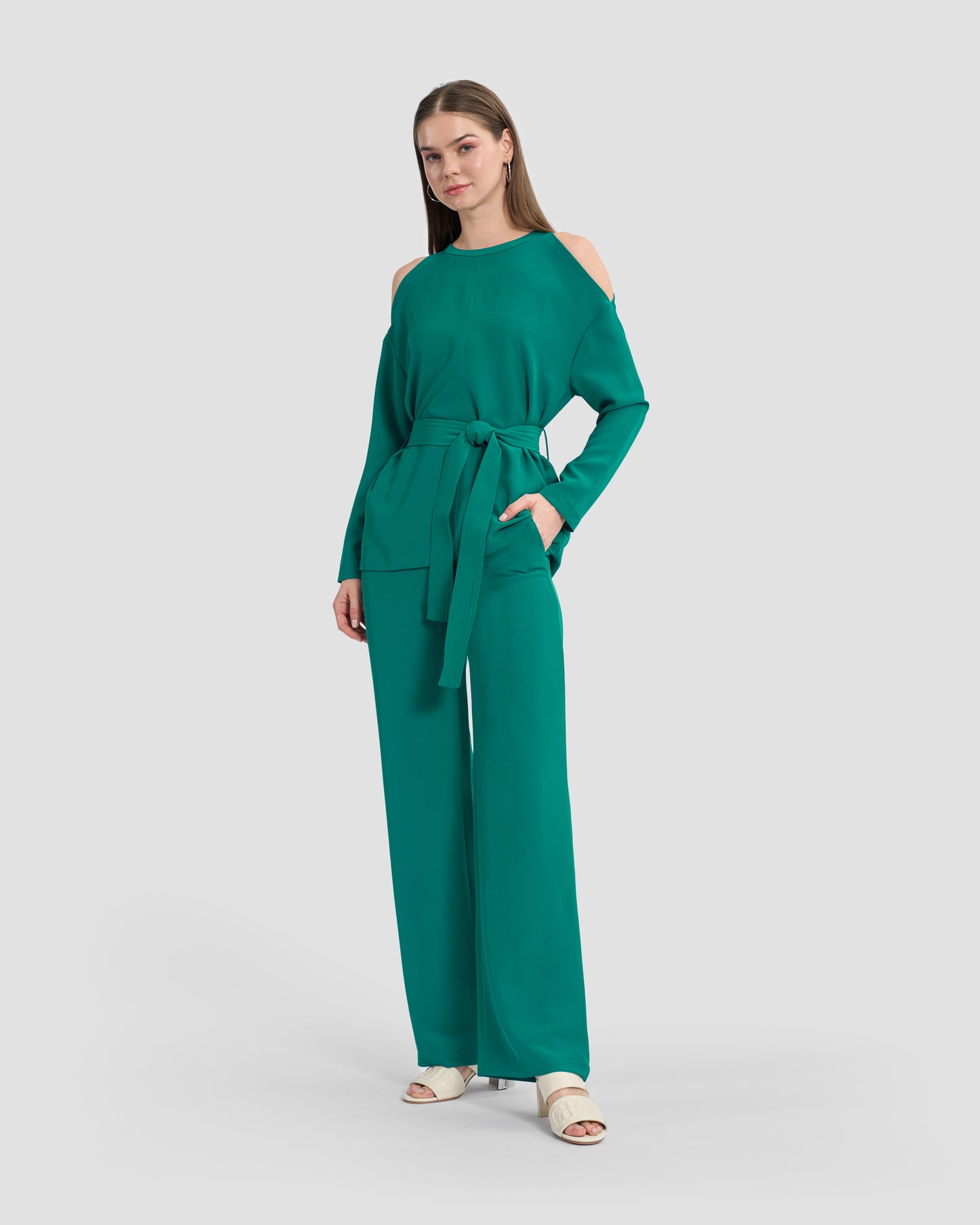 A woman wearing a teal monogram-embroidered cold-shoulder top with a waist sash and a gunmetal GFF embellishment on the hem paired with coordinated trousers and beige sandals.