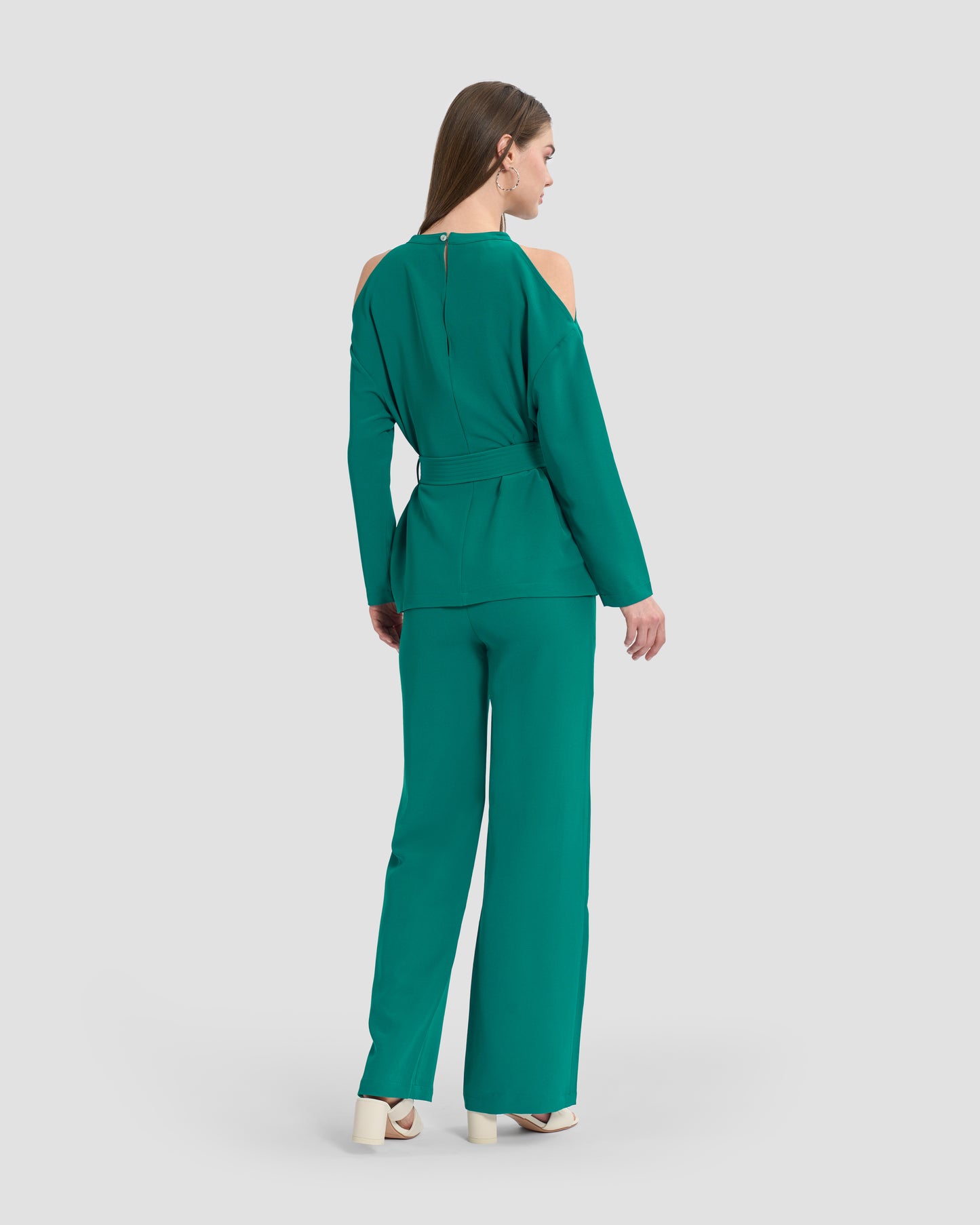 A woman wearing a teal monogram-embroidered cold-shoulder top with a waist sash and a gunmetal GFF embellishment on the hem paired with coordinated trousers and beige sandals.