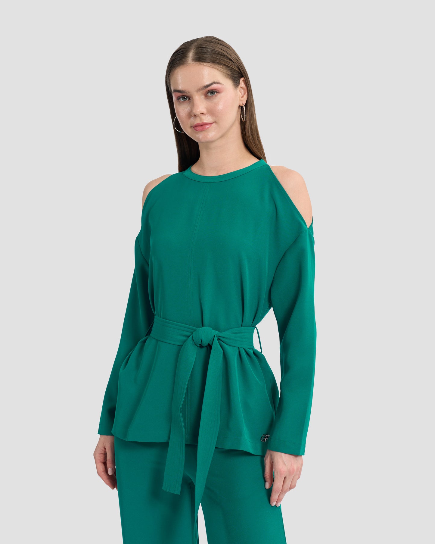 A woman wearing a teal monogram-embroidered cold-shoulder top with a waist sash and a gunmetal GFF embellishment on the hem paired with coordinated trousers.
