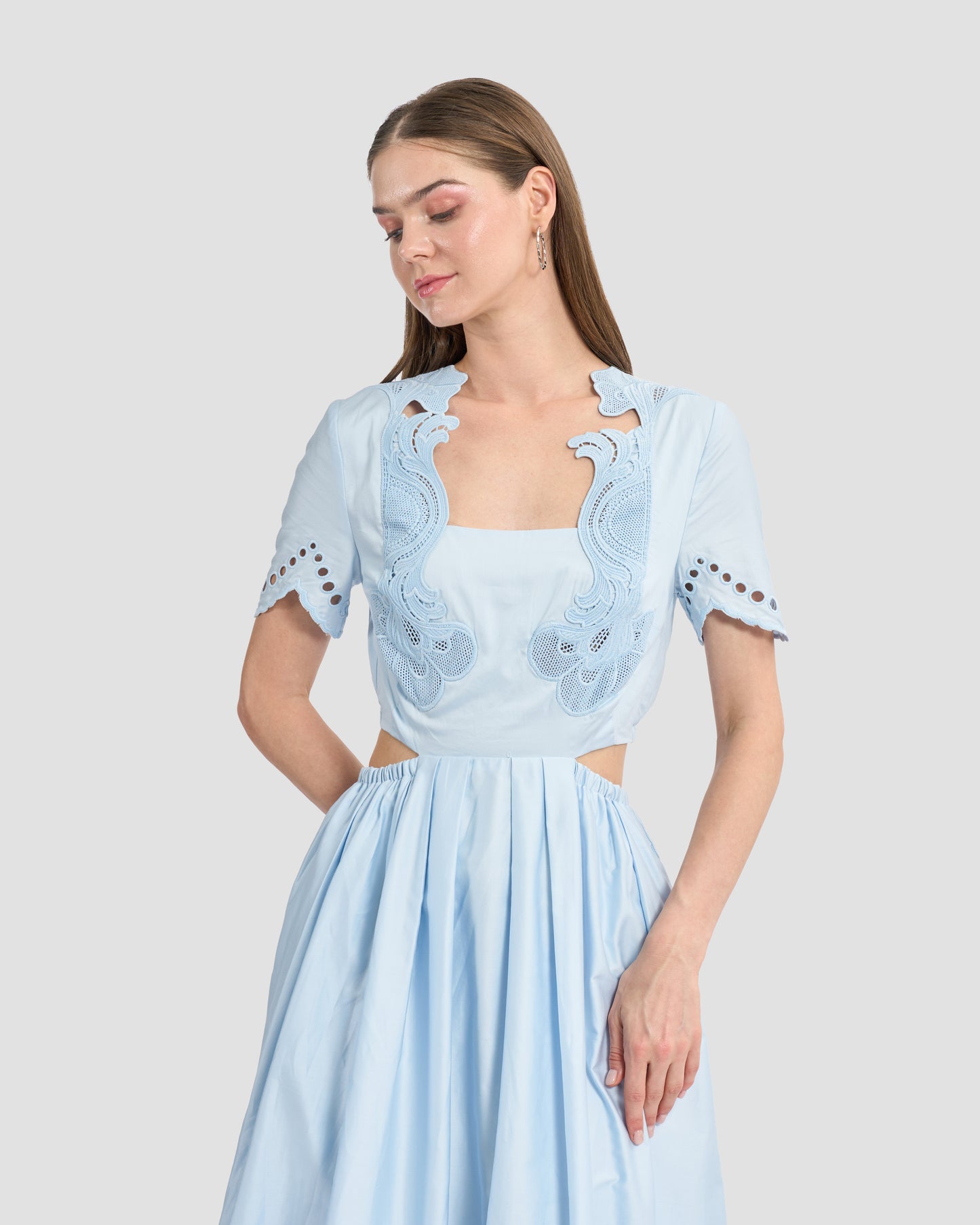 A woman wearing a light blue midi dress with intricate cutout embroidery details on the bodice and sleeves. The dress features a fitted waist and cut-out sides.