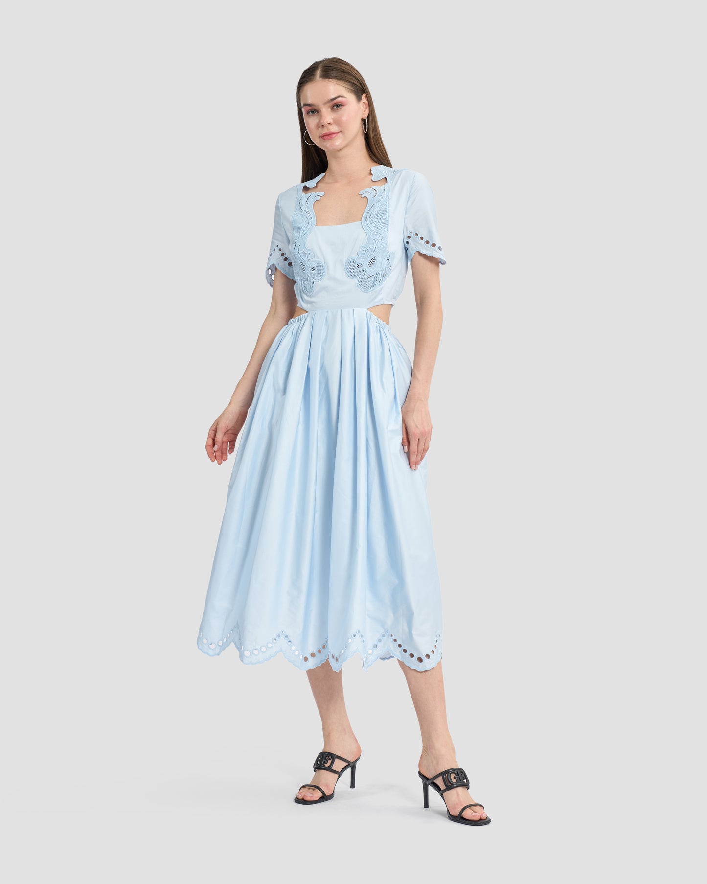 A woman wearing a light blue midi dress with intricate cutout embroidery details on the bodice and sleeves. The dress features a fitted waist, cut-out sides, and a flowy skirt with scalloped hemline, paired with black open-toe heels for a chic and feminine look.
