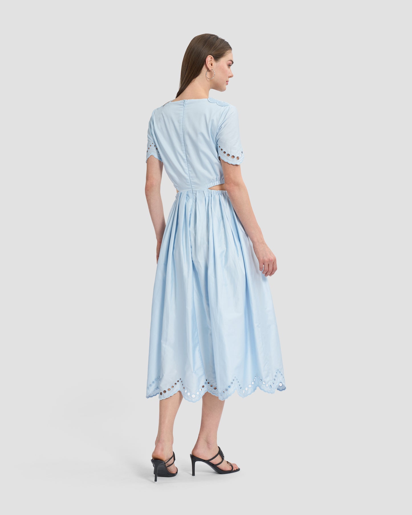 A woman wearing a light blue midi dress with intricate cutout embroidery details on the bodice and sleeves. The dress features a fitted waist, cut-out sides, and a flowy skirt with scalloped hemline, paired with black open-toe heels for a chic and feminine look.