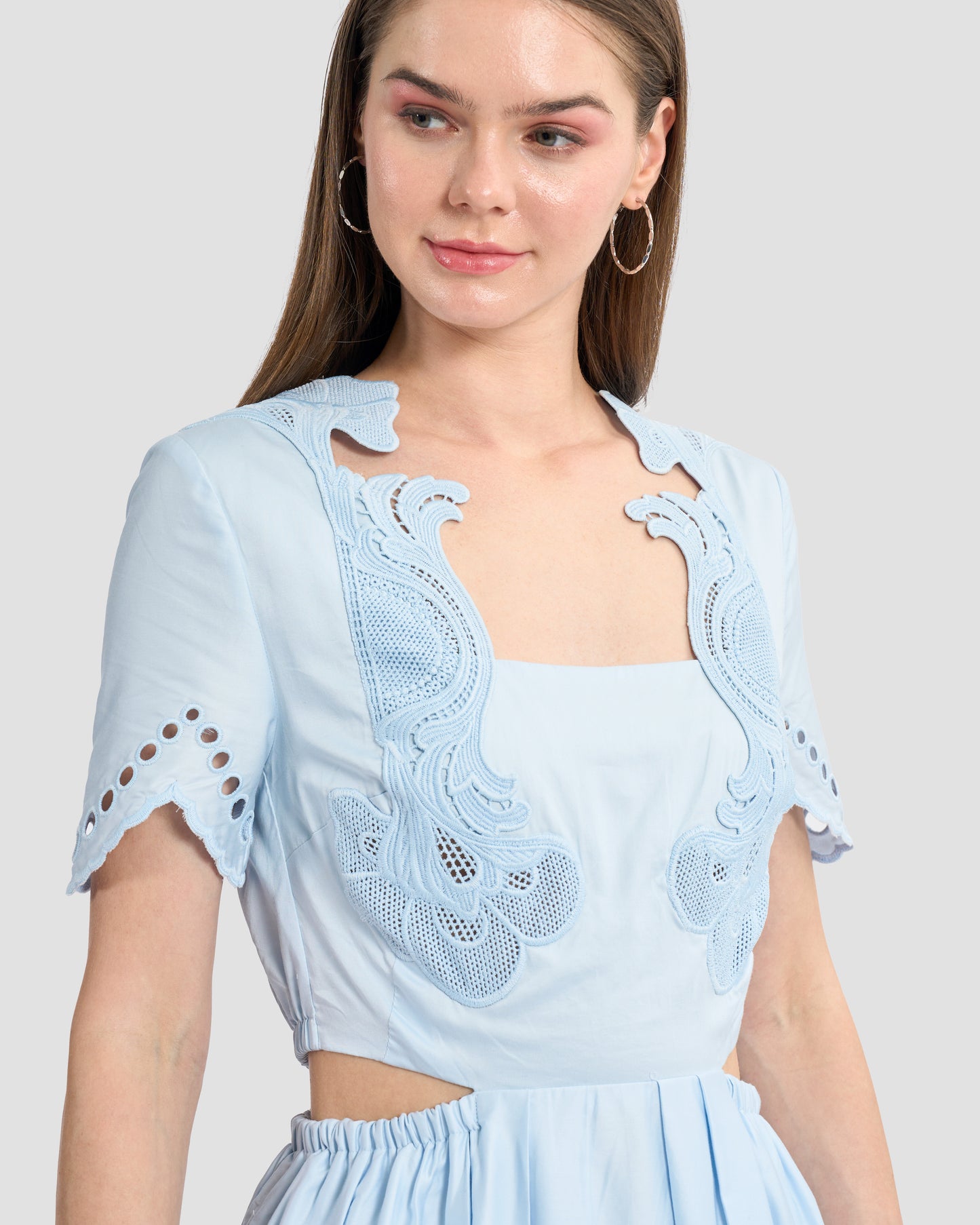 A woman wearing a light blue midi dress with intricate cutout embroidery details on the bodice and sleeves. The dress features a fitted waist and cut-out sides.