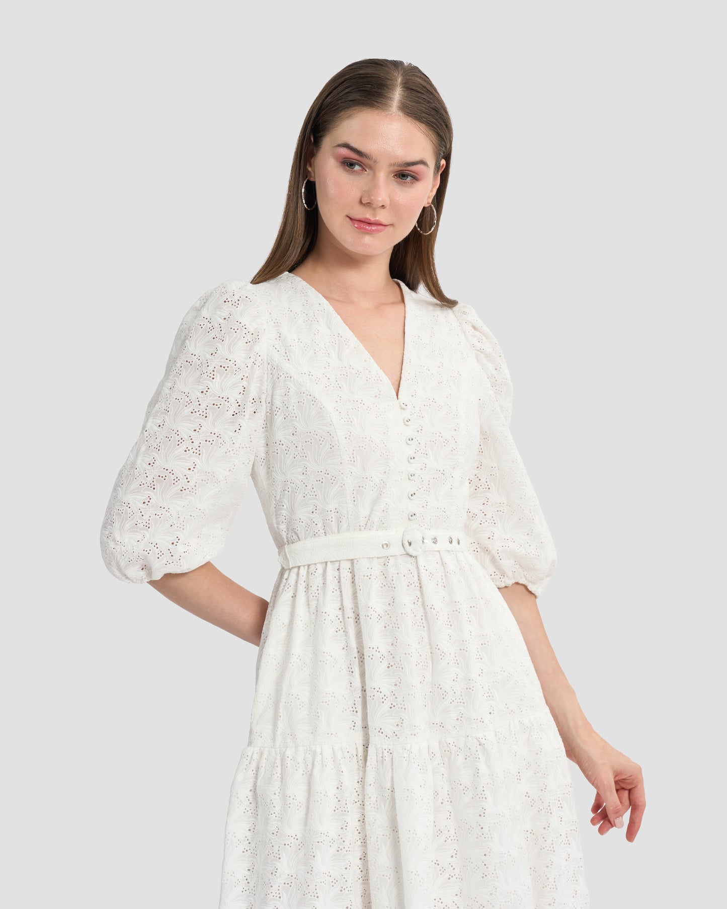 A woman wearing a white, eyelet lace midi dress with puffed three-quarter sleeves and a V-neckline. The dress has a tiered skirt and a fitted waist with button detailing on the front.