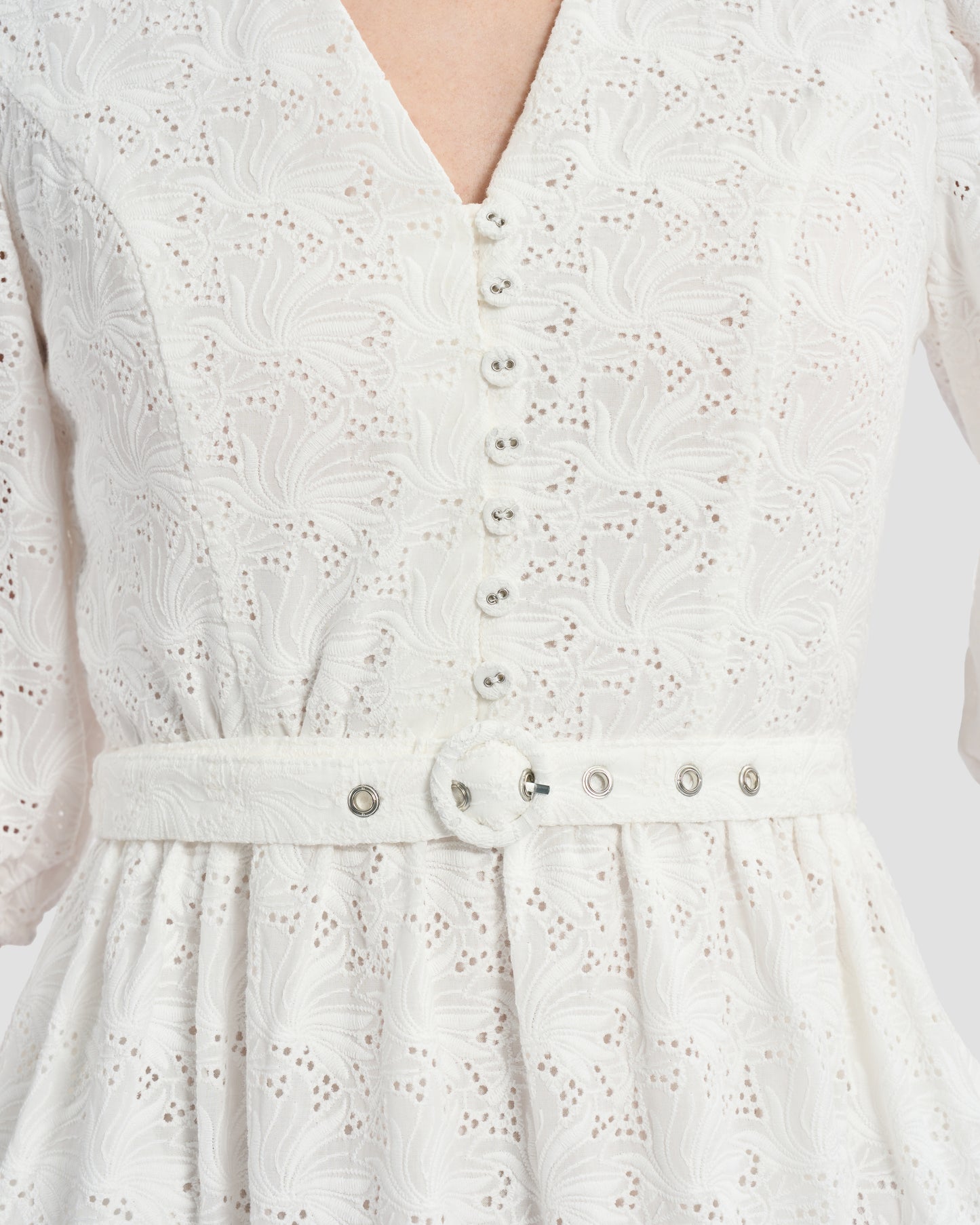 A woman wearing a white, eyelet lace midi dress with puffed three-quarter sleeves. The dress has a fitted waist with button detailing on the front.