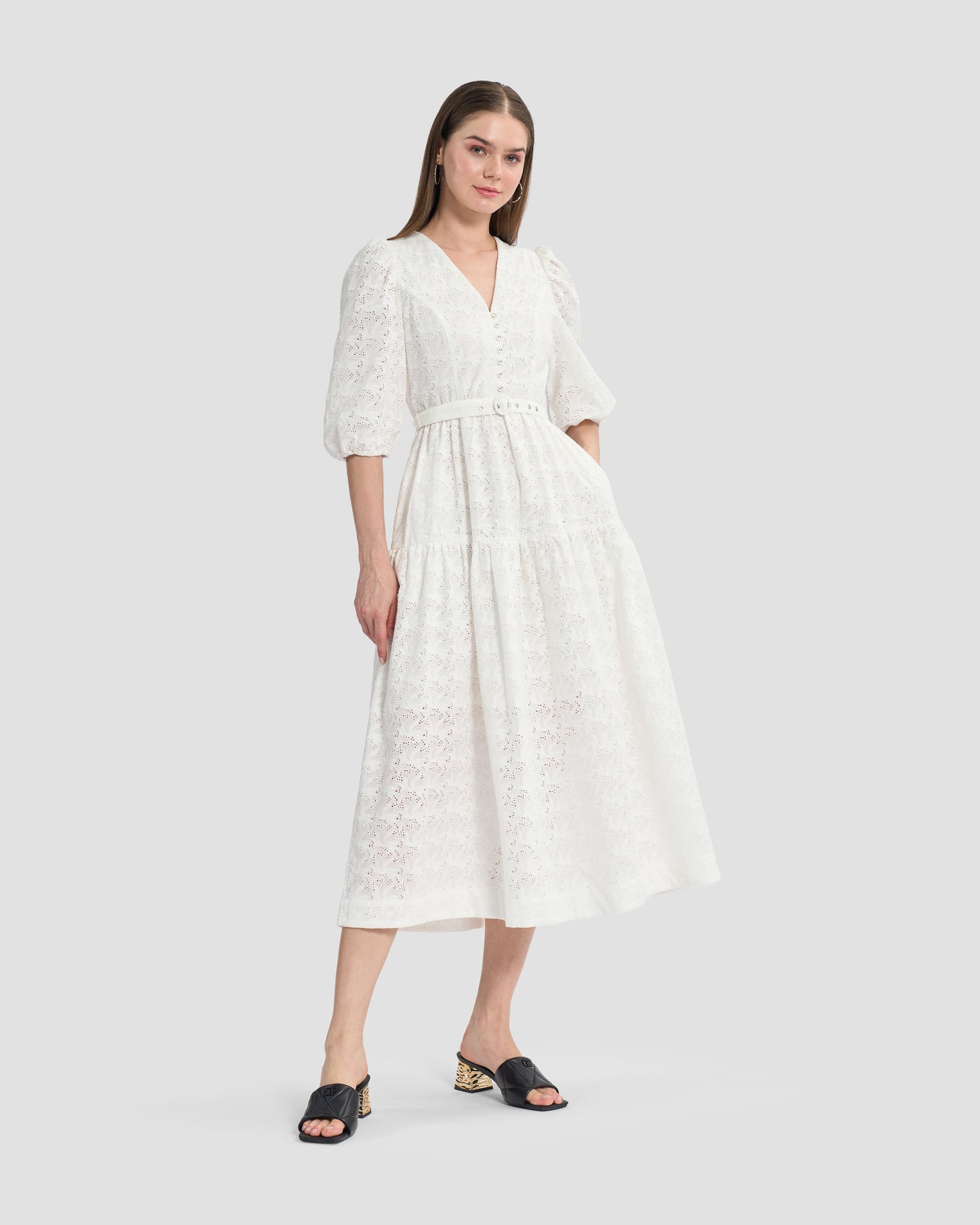 A woman wearing a white, eyelet lace midi dress with puffed three-quarter sleeves and a V-neckline. The dress has a tiered skirt and a fitted waist with button detailing on the front, paired with black open-toe mules featuring gold accents on the heels.
