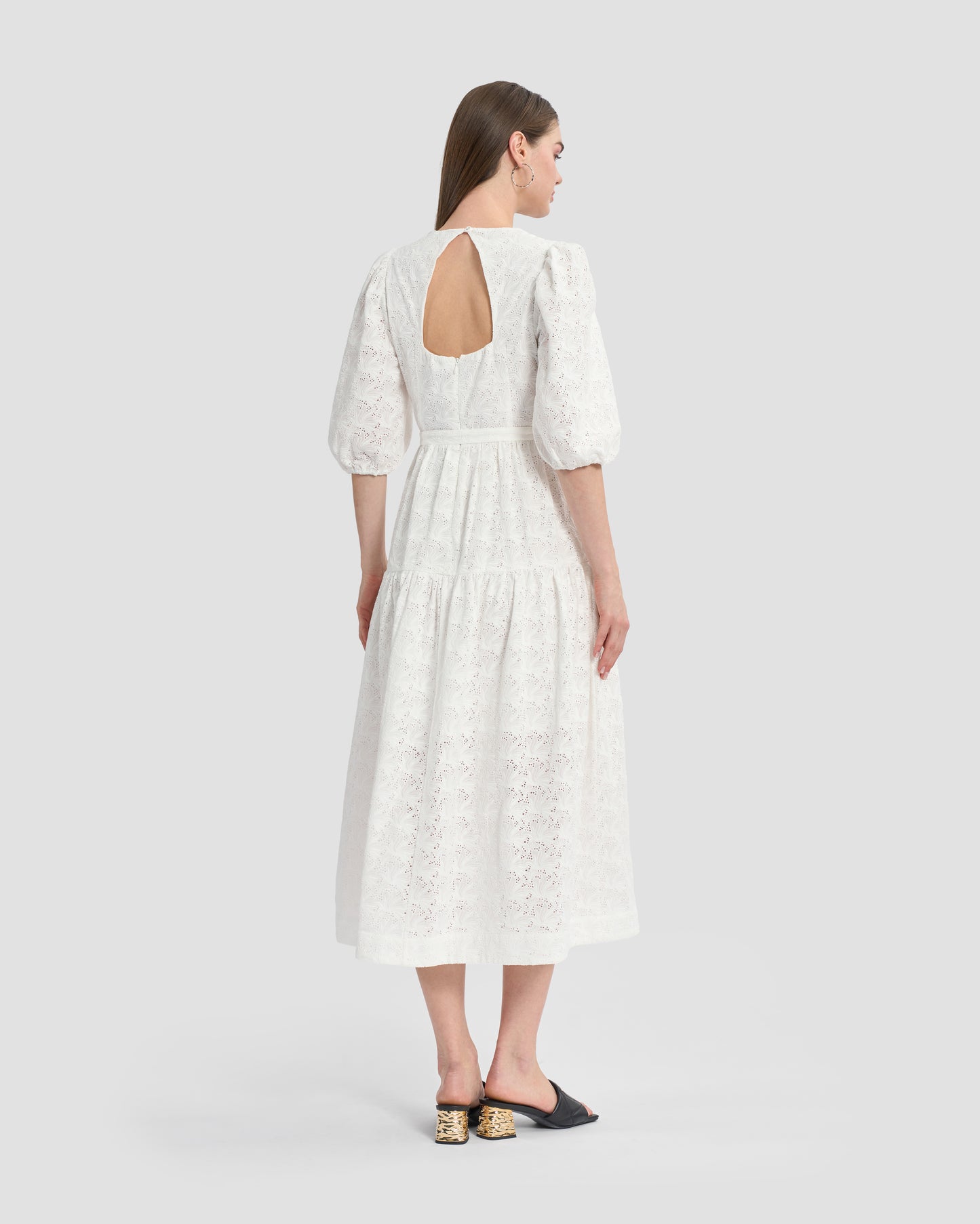 A woman wearing a white, eyelet lace midi dress with puffed three-quarter sleeves and cut-out back. The dress has a tiered skirt and a fitted waist with button detailing on the front, paired with black open-toe mules featuring gold accents on the heels.