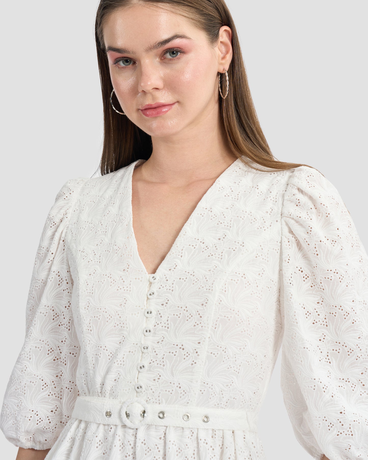 A woman wearing a white, eyelet lace midi dress with puffed three-quarter sleeves and a V-neckline. The dress has a tiered skirt and a fitted waist with button detailing on the front.