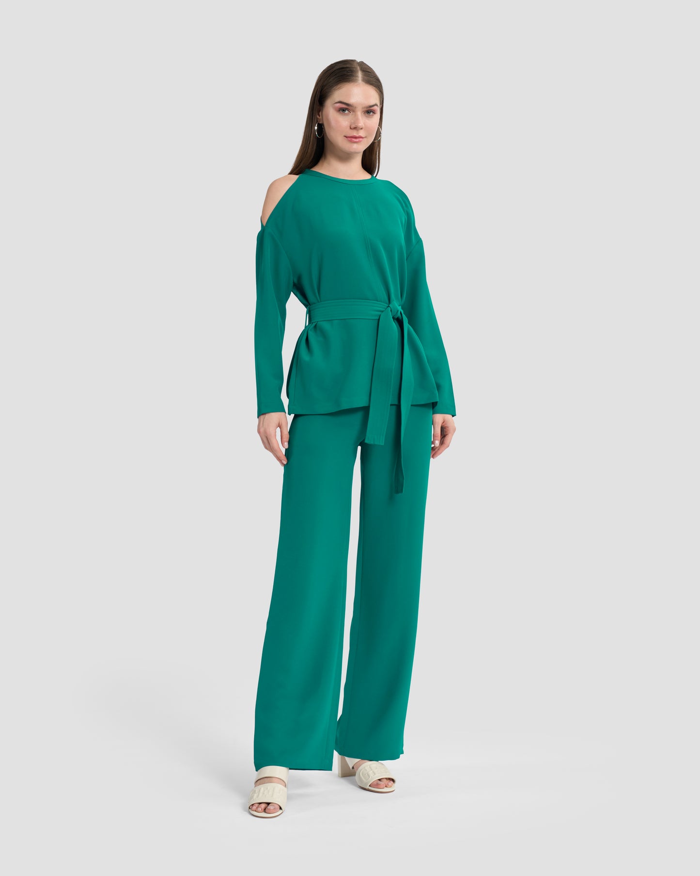 Woman wearing teal flared trousers embellished with a gunmetal monogram at the waistband, paired with a coordinated cold-shoulder top featuring matching monogram embellishments to hem. She completes the look with beige sandals.