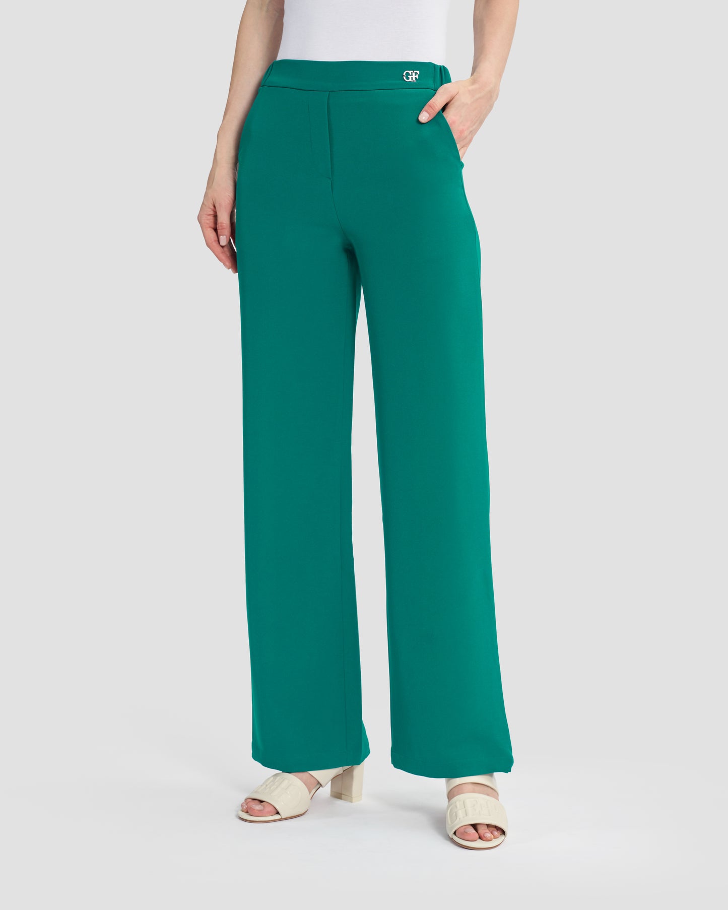 Woman wearing teal flared trousers embellished with a gunmetal monogram at the waistband, paired with beige sandals.