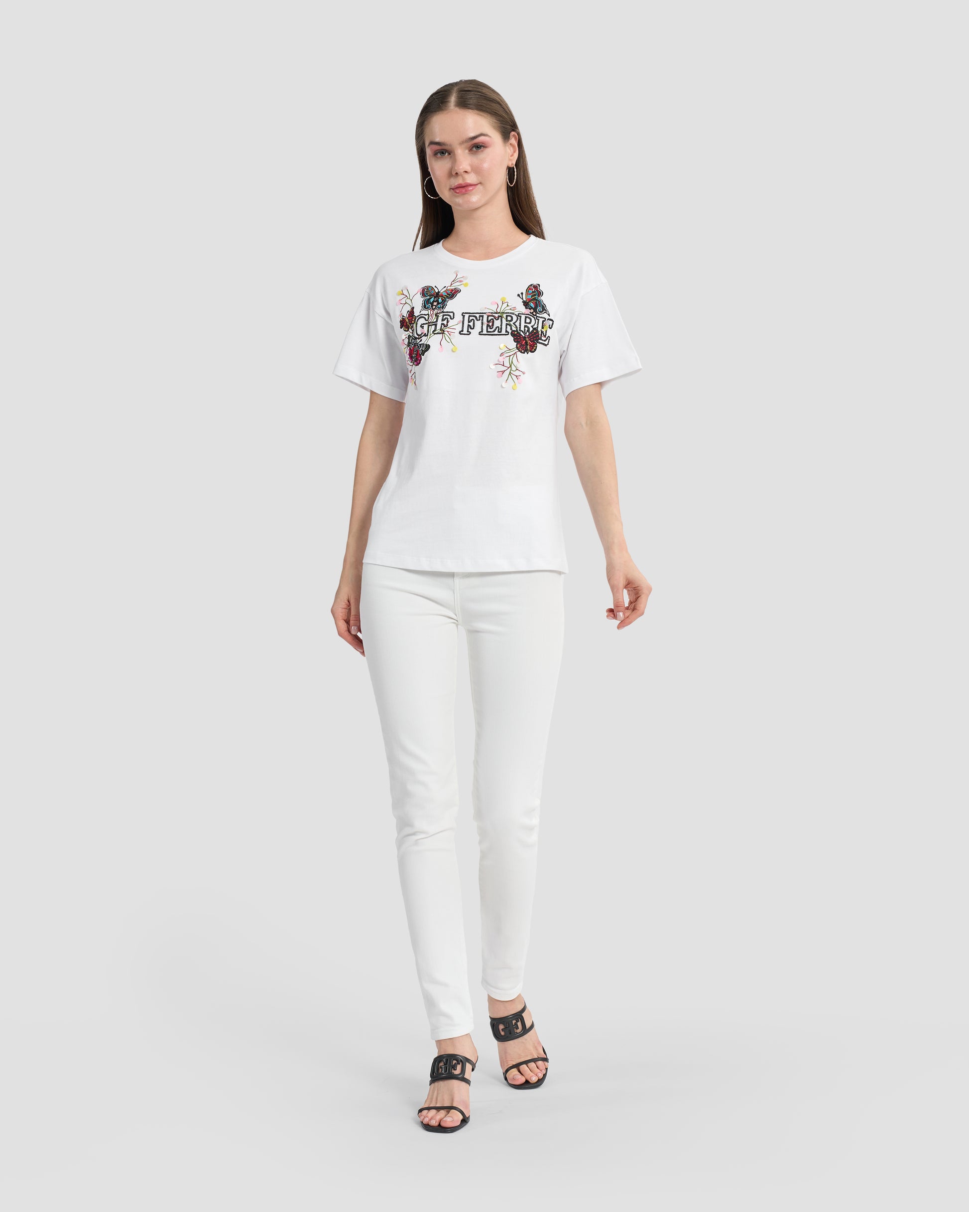 A woman wearing a white t-shirt with butterfly patches to the front, beaded Ferré branding, and multi-toned sequins paired with white jeans and black open-toe heels.