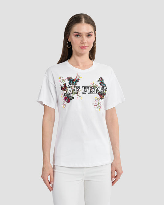 A woman wearing a white t-shirt with butterfly patches to the front, beaded Ferré branding, and multi-toned sequins paired with a white jeans.
