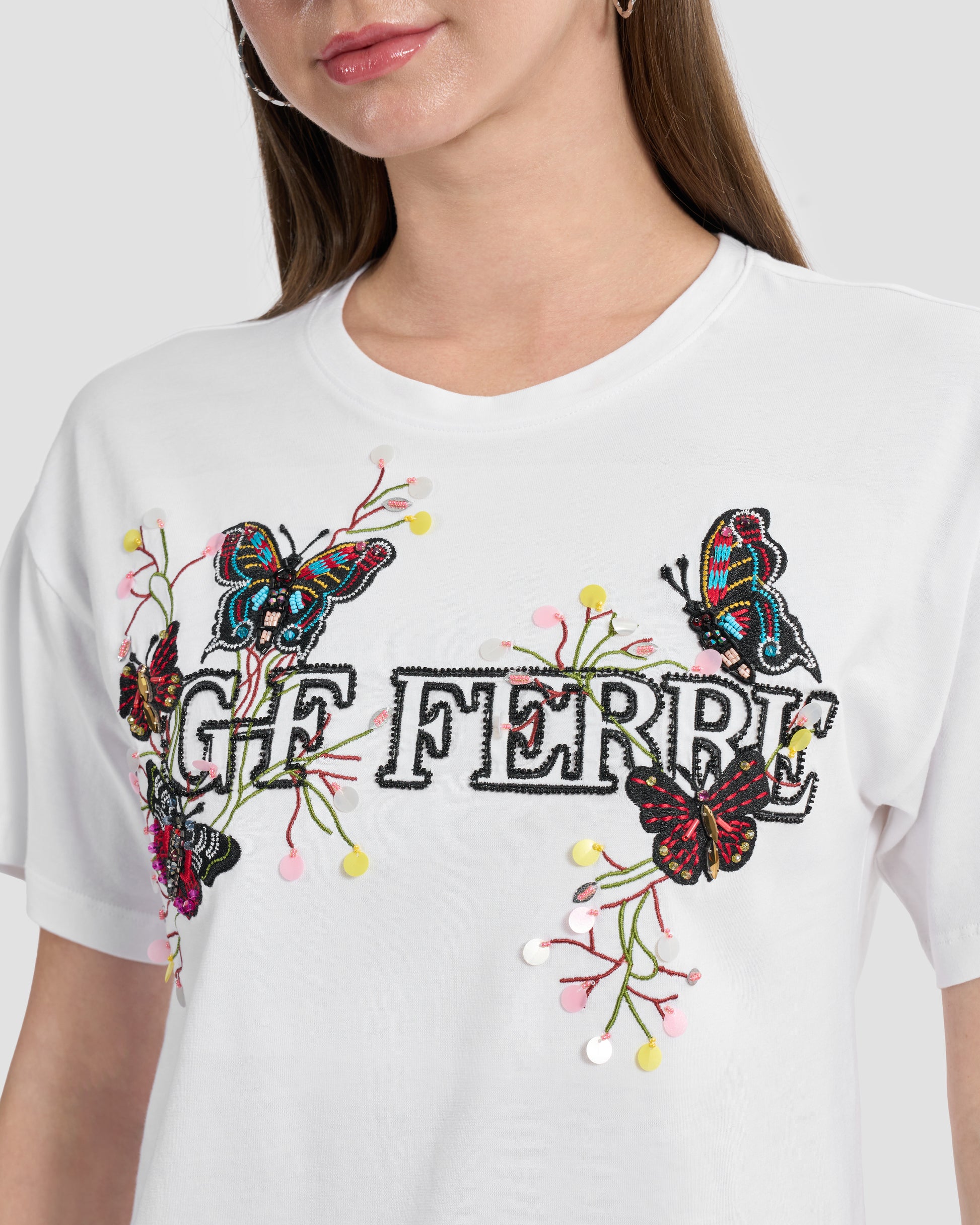 A woman wearing a white t-shirt with butterfly patches to the front, beaded Ferré branding, and multi-toned sequins.
