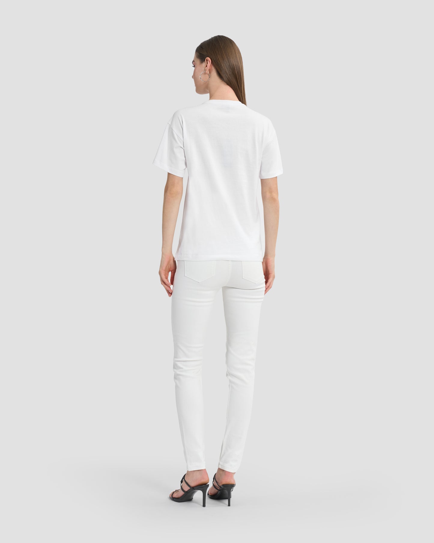 A woman wearing a white t-shirt with butterfly patches to the front, beaded Ferré branding, and multi-toned sequins paired with white jeans and black open-toe heels.
