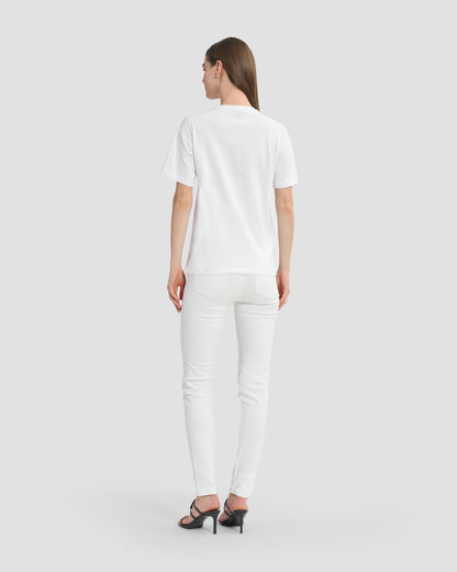 A woman wearing a white t-shirt with butterfly patches to the front, beaded Ferré branding, and multi-toned sequins paired with white jeans and black open-toe heels.
