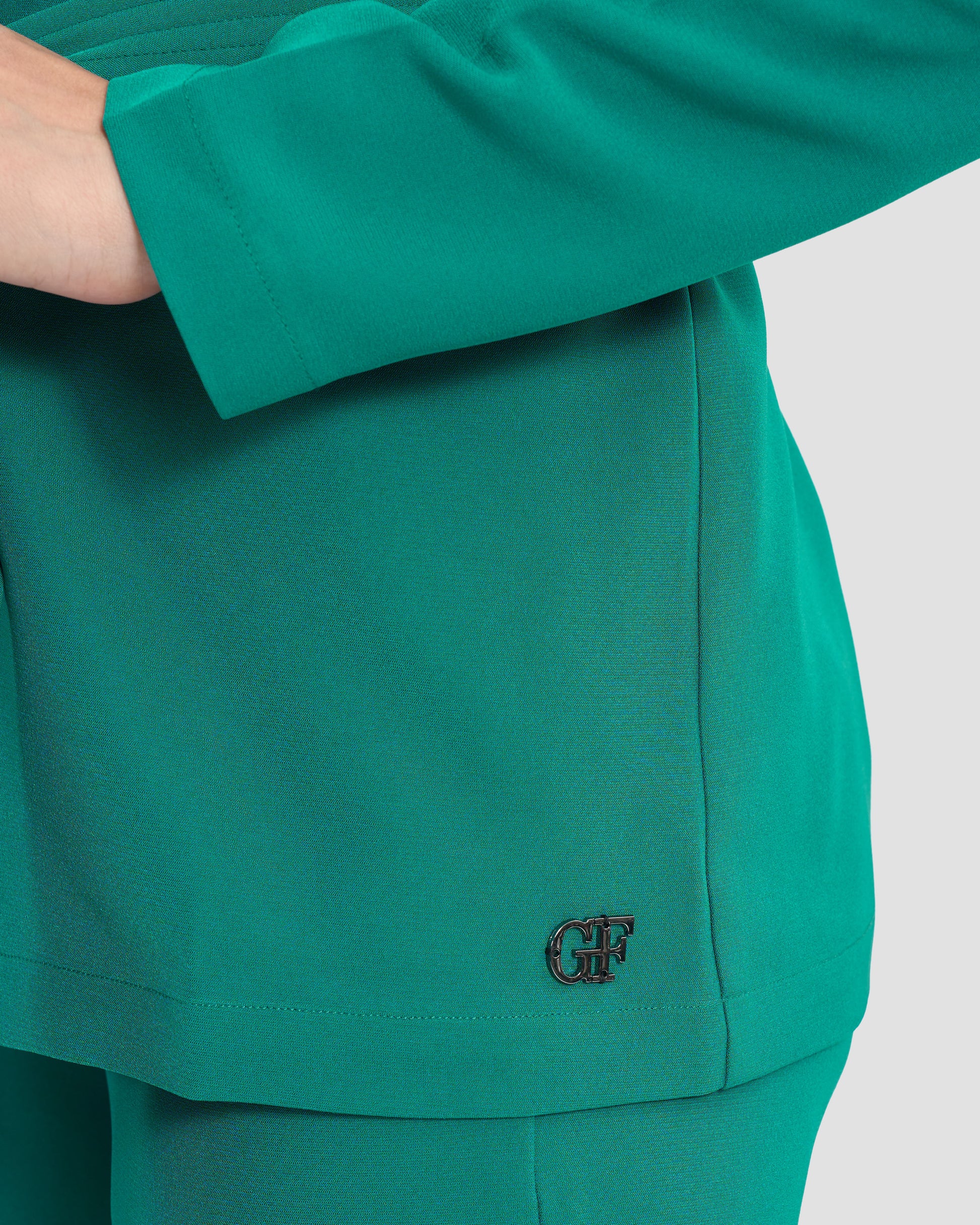 A woman wearing a teal monogram-embroidered cold-shoulder top with a waist sash and a gunmetal GFF embellishment on the hem paired with coordinated trousers.
