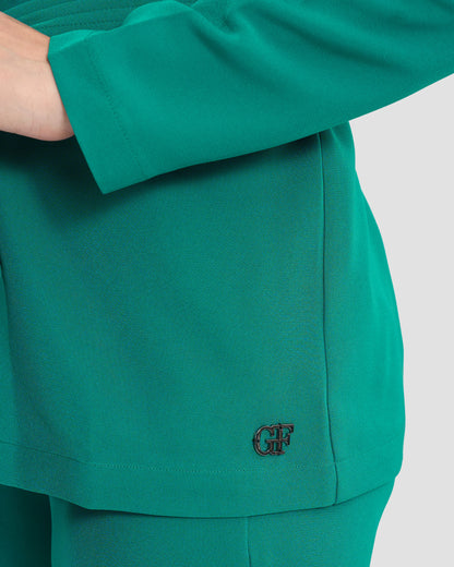 A woman wearing a teal monogram-embroidered cold-shoulder top with a waist sash and a gunmetal GFF embellishment on the hem paired with coordinated trousers.
