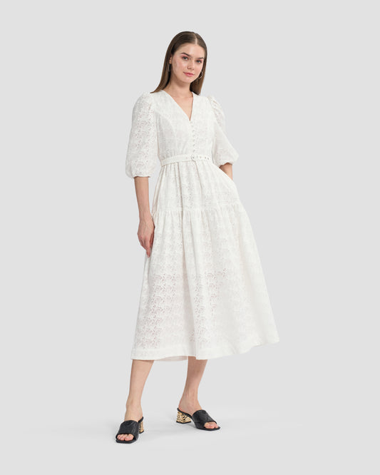 A woman wearing a white, eyelet lace midi dress with puffed three-quarter sleeves and a V-neckline. The dress has a tiered skirt and a fitted waist with button detailing on the front, paired with black open-toe mules featuring gold accents on the heels.